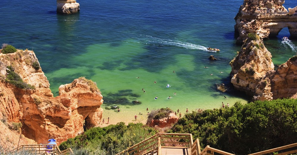 En inbjudande strandvik omgärdad av rödaktiga klippor och grönska, med klart, turkost vatten och människor som badar och solar.