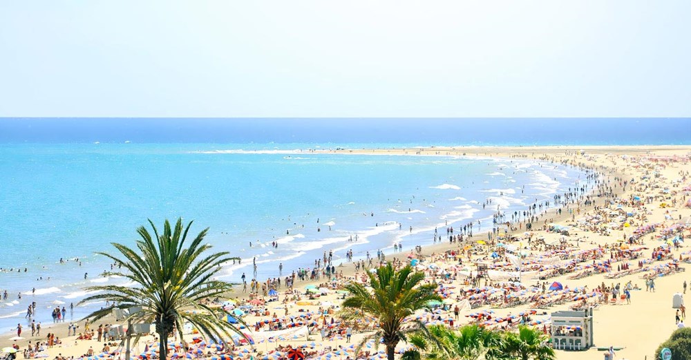 En livlig strand full av människor, med parasoller i olika färger, bredvid ett turkost hav. Två palmer förgrunden till vänster.