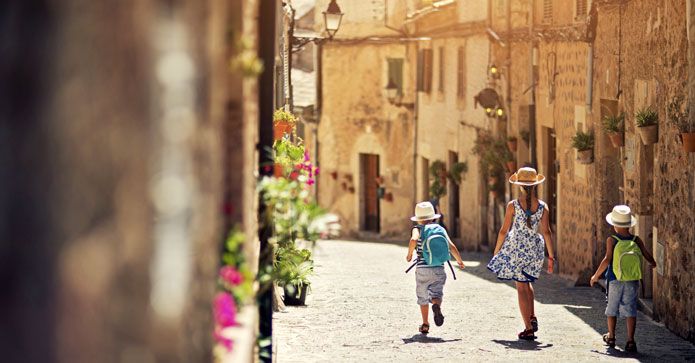 En kvinna och två barn promenerar genom en pittoresk gammal gata med kullerstenar, omgiven av historiska byggnader och blomsterprydda fasader.