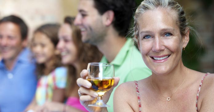 En kvinna i förgrunden ler mot kameran medan hon håller ett glas med dryck. I bakgrunden skymtar ett glatt sällskap av människor som socialiserar.